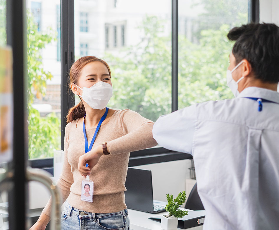 An asian woman and male co-workers bumping elbows. Both are wearing masks.