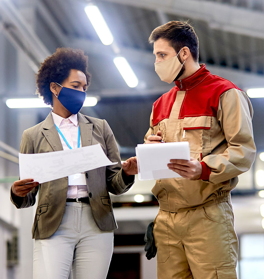A manager woman of color chatting with a male coworker in a warehouse. Both are wearing masks.