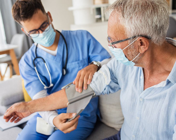Male nurse measures blood pressure to senior man with mask while being in a home visit